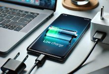 "A close-up image of a smartphone with a visible battery indicator showing a low charge. The phone is on a table next to a charger, with a power cord plugged in. The background features a modern tech environment with a laptop and a cup of coffee."
