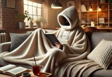 A person lounging on a couch wearing a cozy, oversized wearable blanket with a hood, reading a book and sipping tea. The room has soft lighting and a warm ambiance, highlighting the comfort and practicality of the wearable blanket in a relaxing environment.